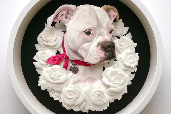 A paper-cut portrait of a white dog surrounded by a wreath of white paper roses and hosued in a circular frame.