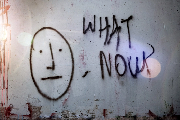 Spray-painted face on a wall next to the words 'What Now?'.